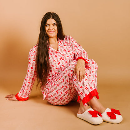 A woman with long dark hair sits on a tan background, wearing The Darling Effect's Oh So Merry Sweet Dreams Pajama Pants Set and white slippers adorned with red bows.