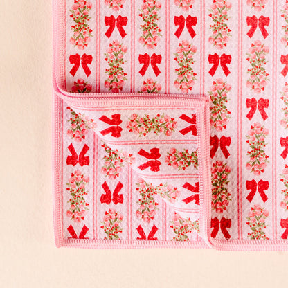 The Darling Effect’s Oh So Merry Waffle Tea Towel features a soft pink design with red bows and small floral bouquets, shown with one corner folded to reveal both sides of its absorbent fabric.