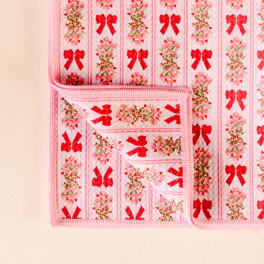 The Darling Effect’s Oh So Merry Waffle Tea Towel features a soft pink design with red bows and small floral bouquets, shown with one corner folded to reveal both sides of its absorbent fabric.