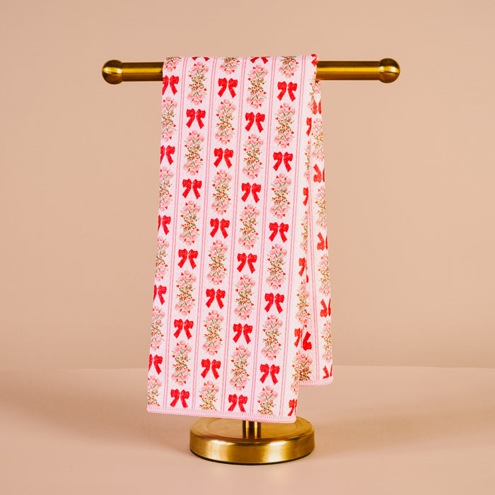 The Darling Effect's Oh So Merry Waffle Tea Towel, featuring a red bow and floral pattern, hangs neatly on a gold stand against a beige background—an absorbent and charming kitchen accessory.