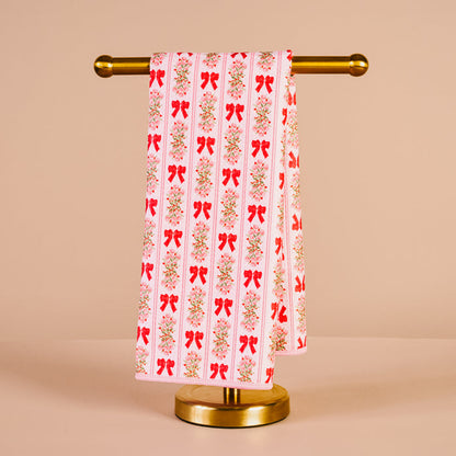 The Darling Effect's Oh So Merry Waffle Tea Towel, featuring a red bow and floral pattern, hangs neatly on a gold stand against a beige background—an absorbent and charming kitchen accessory.