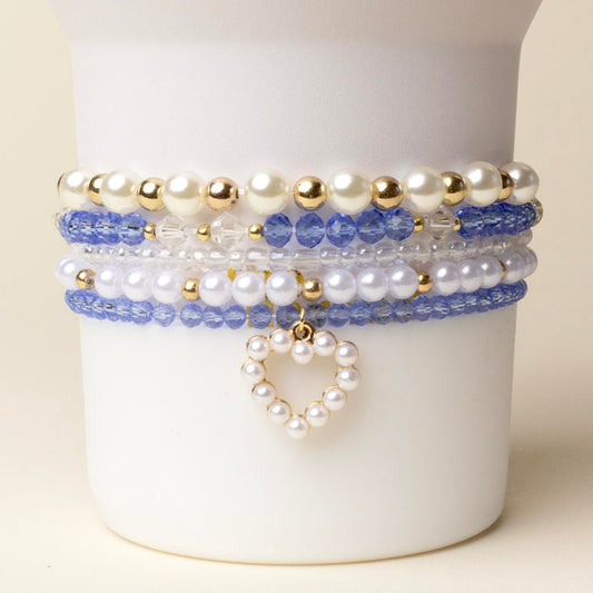 The On Cloud Nine Tumbler Bracelet Stack by the-darling-effect features white pearls, gold beads, blue and clear crystals, plus a heart charm—displayed on a white stand. Perfect accessory for your 40 oz tumbler!.