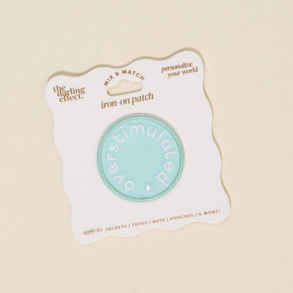 The Darling Effect’s Overstimulated Mix & Match Iron-On Patch features "overstimulated" in white on a teal circle, mounted on a white card—ideal for customizing your style or adding to your colorful patch collection.