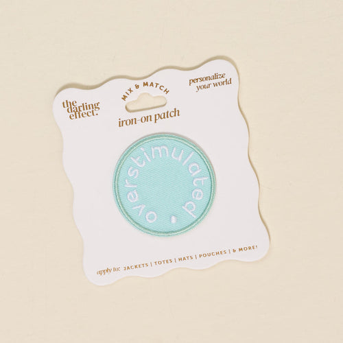 The Darling Effect’s Overstimulated Mix & Match Iron-On Patch features "overstimulated" in white on a teal circle, mounted on a white card—ideal for customizing your style or adding to your colorful patch collection.