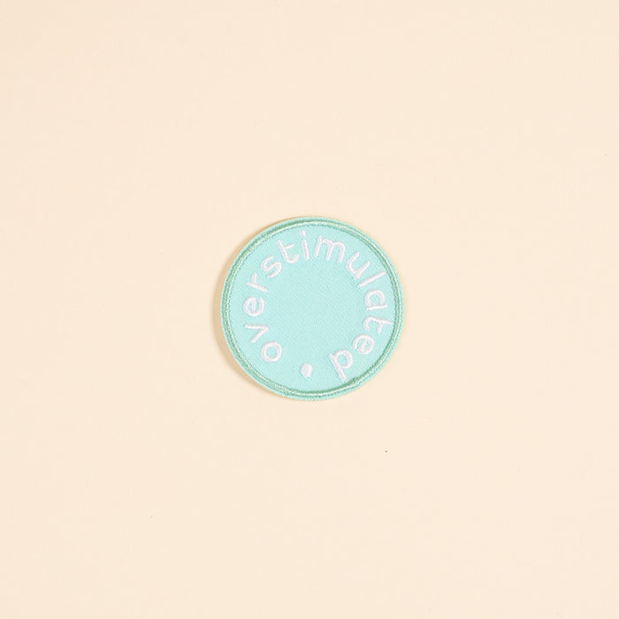 The Darling Effect's Overstimulated Mix & Match Iron-On Patch features a circular teal design with white "overstimulated" text embroidered around the edge. Personalize it with your favorite designs for a unique look.