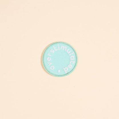 The Darling Effect's Overstimulated Mix & Match Iron-On Patch features a circular teal design with white "overstimulated" text embroidered around the edge. Personalize it with your favorite designs for a unique look.