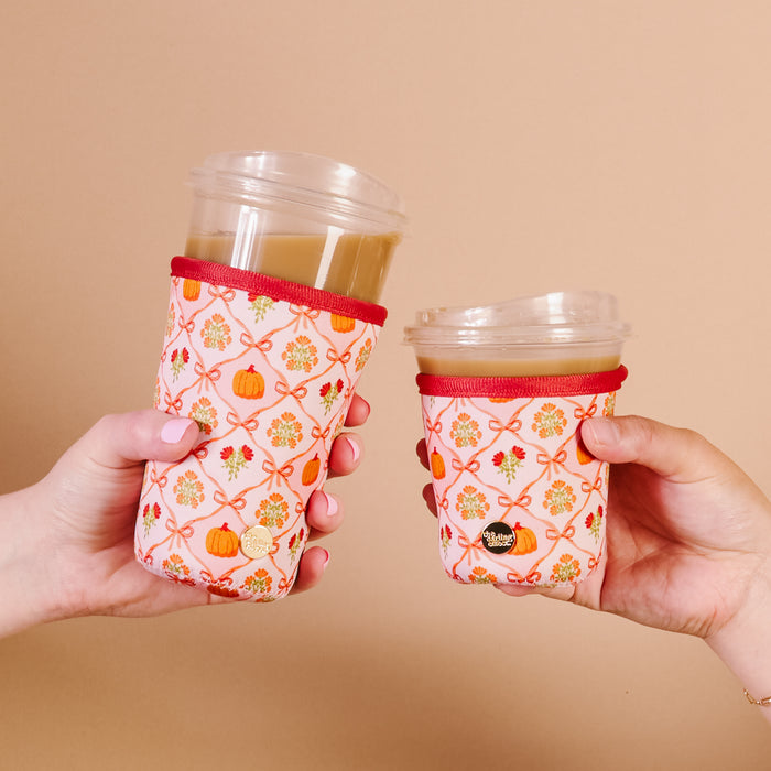 Two hands hold clear plastic coffee cups, each with The Darling Effect’s Patchwork Pumpkins Sip Savvy Drink Sleeve—pink neoprene sleeves decorated with orange bows, small flowers, and pumpkins for a festive touch.