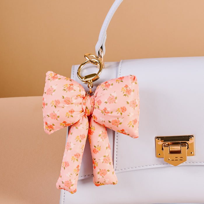 A white handbag with a gold clasp is styled with The Darling Effect’s Peachy Petals Puffy Bow Bag Charm, a coquette-inspired accessory featuring small floral prints and a gold keyring. The background is beige.