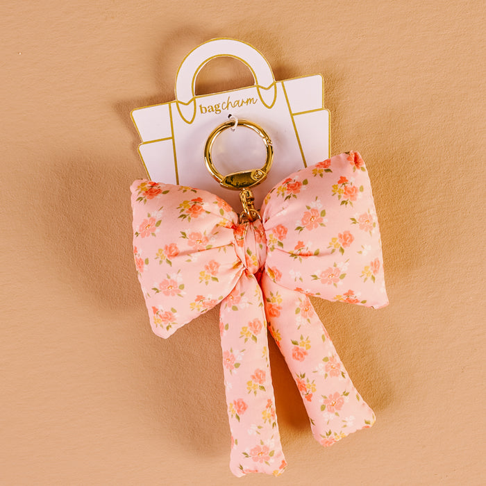 The Darling Effect’s Peachy Petals Puffy Bow Bag Charm features a floral pink bow on a gold keyring, displayed on a white "bagcharm" card against a beige background.