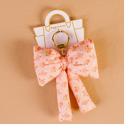 The Darling Effect’s Peachy Petals Puffy Bow Bag Charm features a floral pink bow on a gold keyring, displayed on a white "bagcharm" card against a beige background.
