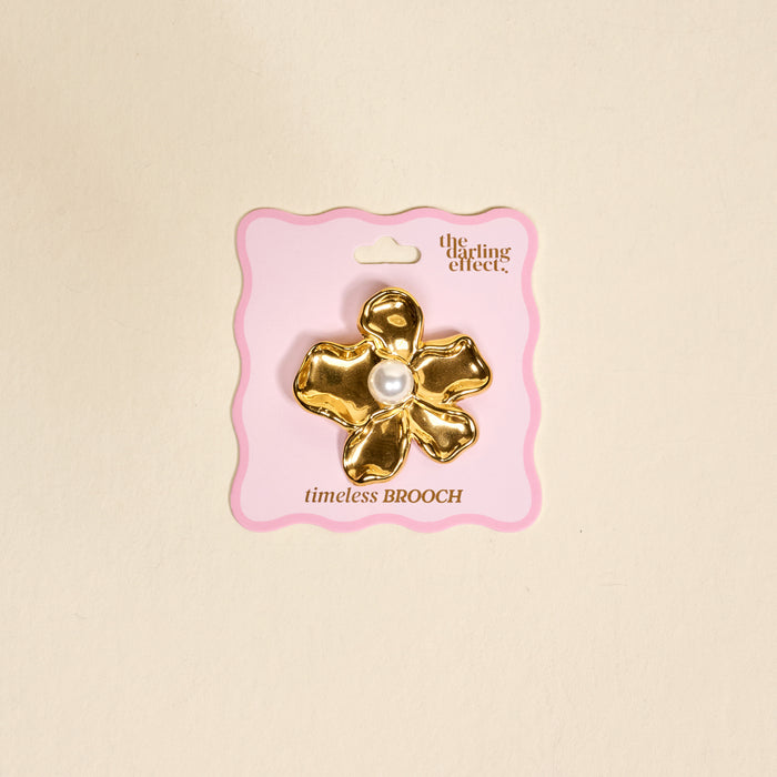 The Pearl Core Flower Timeless Brooch by The Darling Effect features a gold vintage-inspired design with a large pearl center, displayed on pink packaging and set against a beige background—an elegant keepsake accessory.