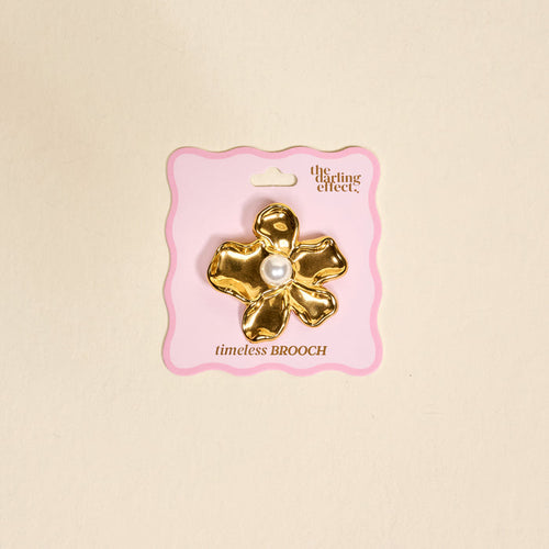 The Pearl Core Flower Timeless Brooch by The Darling Effect features a gold vintage-inspired design with a large pearl center, displayed on pink packaging and set against a beige background—an elegant keepsake accessory.