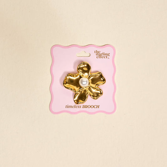 The Pearl Core Flower Timeless Brooch by The Darling Effect features a gold vintage-inspired design with a large pearl center, displayed on pink packaging and set against a beige background—an elegant keepsake accessory.