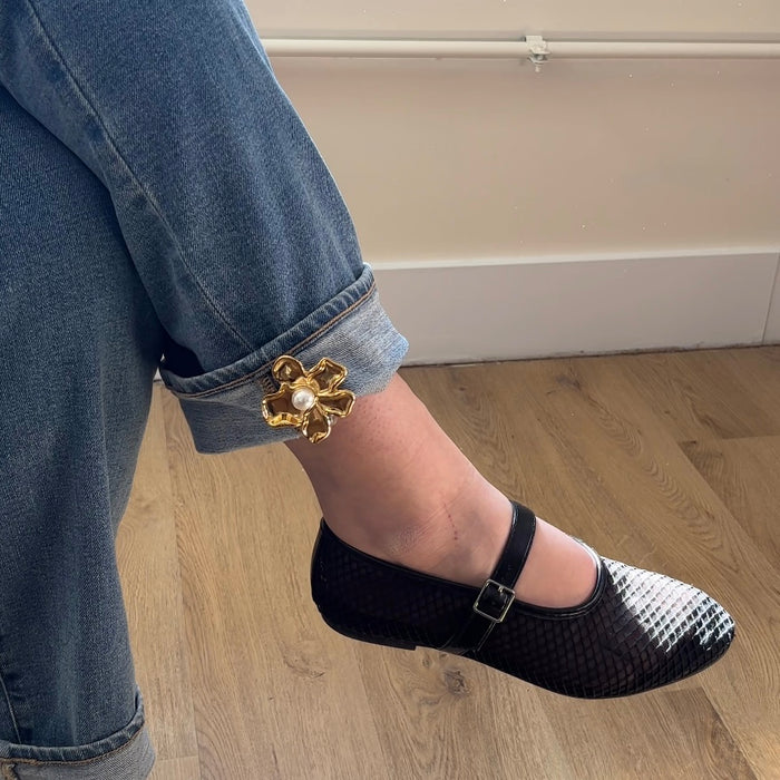 A person in blue jeans with The Darling Effect’s Pearl Core Flower Timeless Brooch on the cuff and black-and-white textured Mary Jane shoes sits on a light wooden floor.