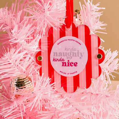 The Darling Effect's Peppermint Stripes Scalloped Round Picture Frame Ornament, featuring pink and white stripes and "kinda naughty, kinda nice" text, hangs on a pink Christmas tree with silver accents. Fits a 2-inch photo.