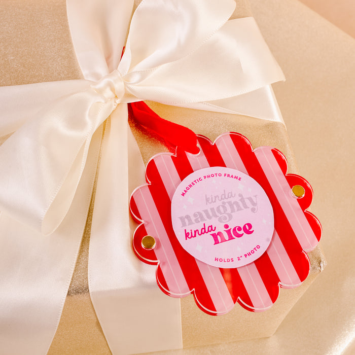 A gold gift box with a white satin ribbon and a red and pink "kinda naughty kinda nice" tag is perfect for gifting The Darling Effect’s Peppermint Stripes Scalloped Round Picture Frame Ornament as a thoughtful holiday surprise.