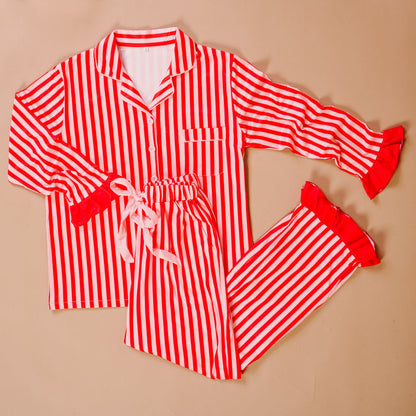 The Darling Effect’s Peppermint Stripes Sweet Dreams Pajama Pants Set features red and white stripes, a button-up collared top with ruffle trim, and drawstring pants—perfectly cozy for relaxing nights. Laid flat on a beige background.