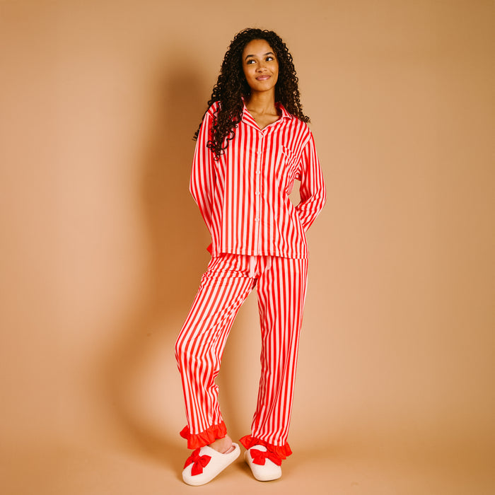A woman with curly hair smiles in The Darling Effect's Peppermint Stripes Sweet Dreams Pajama Pants Set—red-and-white stripes, ultra-soft fabric—paired with white slippers with red bows, standing against a beige background.
