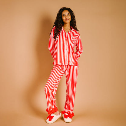 A woman with curly hair smiles in The Darling Effect's Peppermint Stripes Sweet Dreams Pajama Pants Set—red-and-white stripes, ultra-soft fabric—paired with white slippers with red bows, standing against a beige background.