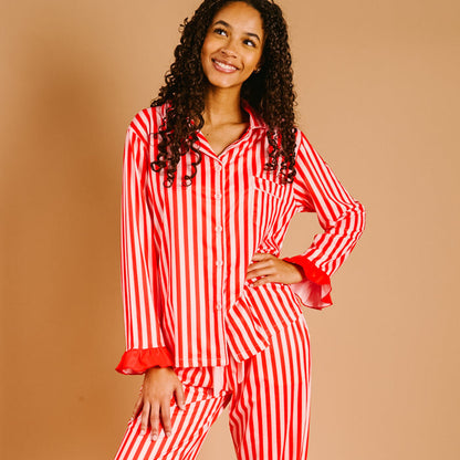 A woman smiles in The Darling Effect's Peppermint Stripes Sweet Dreams Pajama Pants Set featuring red and white vertical stripes and ruffled cuffs, made from ultra-soft fabric, against a light brown background.
