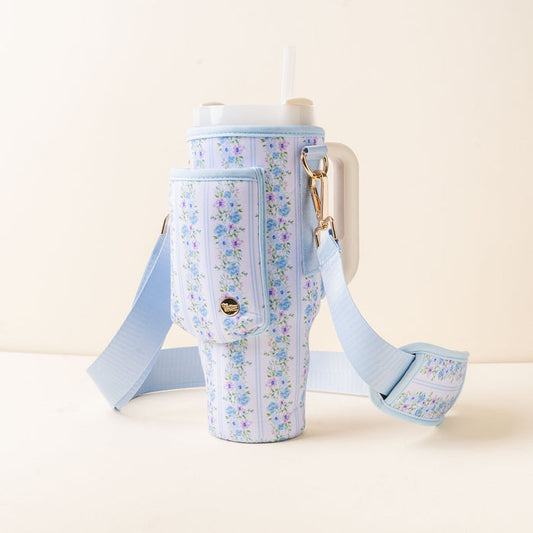 A tall cup with a lid and straw rests in the-darling-effect's Petal Parade Blue Tumbler Sling—a light blue floral carrier with an adjustable strap and front pocket—providing a stylish, hands-free way to carry your drink against a cream background.