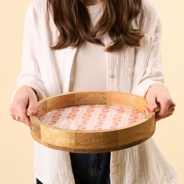 A person in a white shirt holds the-darling-effect Petal Parade Pink Large Round Wood Enamel Tray, featuring a pink floral and striped pattern, against a light beige background.