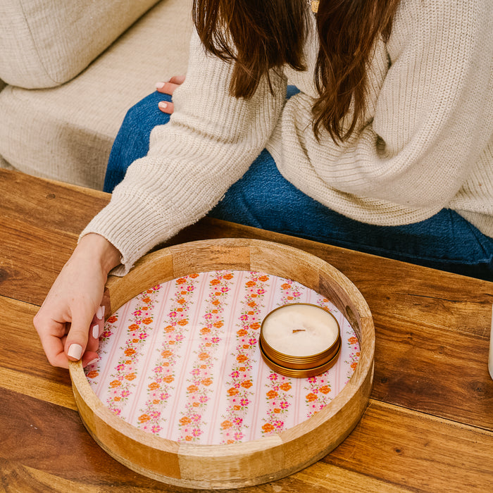 Wearing a white sweater and blue pants, a person sets the-darling-effect’s Petal Parade Pink Large Round Wood Enamel Tray with a candle on a wooden table.