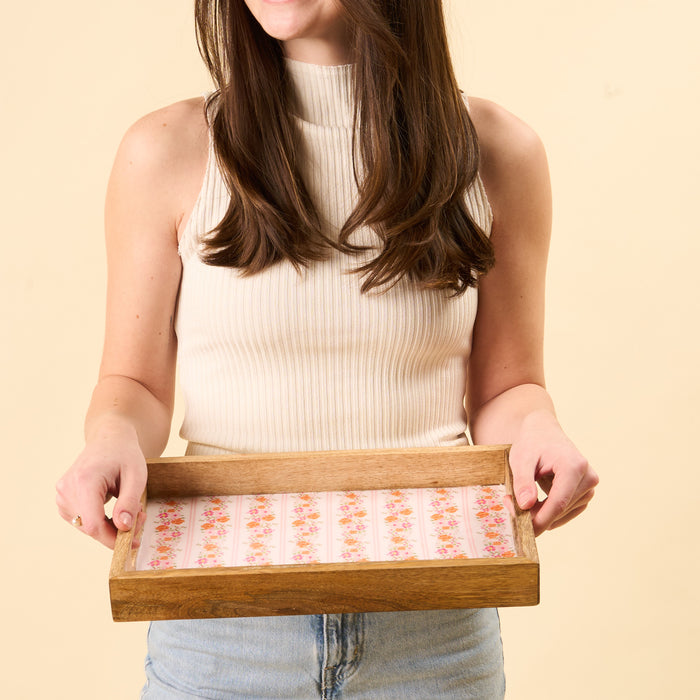 A woman in a sleeveless ribbed top and denim shorts holds the-darling-effect Petal Parade Pink Rectangle Wood Enamel Tray with a floral pattern against a light beige background. Her face is partially out of frame.