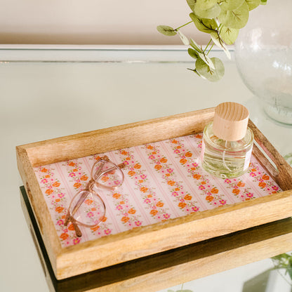 The Petal Parade Pink Rectangle Wood Enamel Tray by the-darling-effect, featuring a floral pattern, holds eyeglasses and a small glass bottle on a reflective surface beside a vase with green leaves.