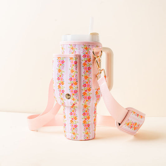 The Petal Parade Pink Tumbler Sling by the-darling-effect holds a large pink tumbler with a handle, white lid, and straw, featuring pastel pink and colorful floral designs, an adjustable strap, and a small pocket for hands-free use.