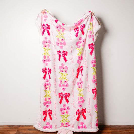 The Darling Effect's Phi Mu Plush Sherpa Blanket, featuring pink and white floral patterns with red bows, hangs against a white wall and drapes over a wooden floor.