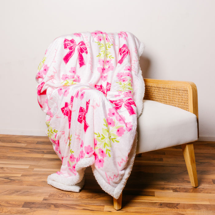 The Darling Effect's Phi Mu Plush Sherpa Blanket features soft white plush with floral patterns and a pink bow, draped over a light wood cane accent chair—perfect as a cozy sorority blanket or stylish college dorm throw.