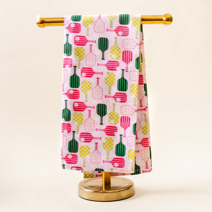 The Pickleball Party Tea Towel by the-darling-effect, featuring vibrant pink, green, and yellow pickleball paddle patterns on white cotton, hangs over a gold stand against a simple light background.
