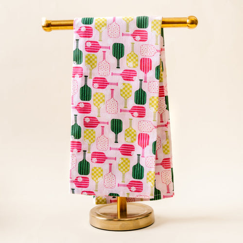 The Pickleball Party Tea Towel by the-darling-effect, featuring vibrant pink, green, and yellow pickleball paddle patterns on white cotton, hangs over a gold stand against a simple light background.