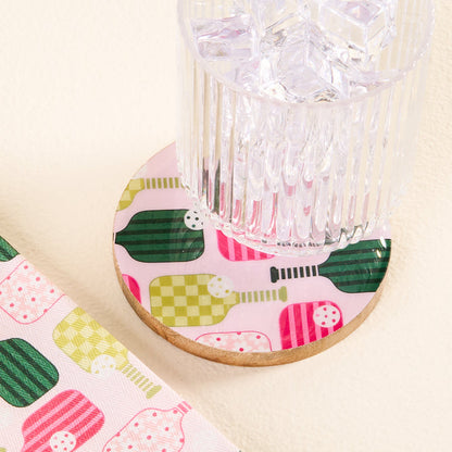 The-darling-effect Pickleball Party Wood Enamel Coaster features a green, pink, and yellow pickleball paddle pattern. Topped with a ribbed glass of ice, it's paired with a matching napkin for a stylish decorative touch.