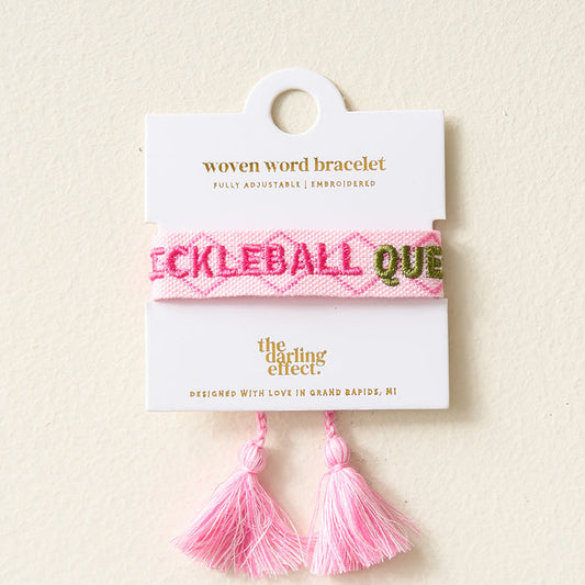 The Darling Effect's Pickleball Queen Woven Bracelet features pink embroidery and tassels, is fully adjustable, and comes on a white card. Designed in Grand Rapids, MI—perfect for fans of trendy embroidered bracelets.