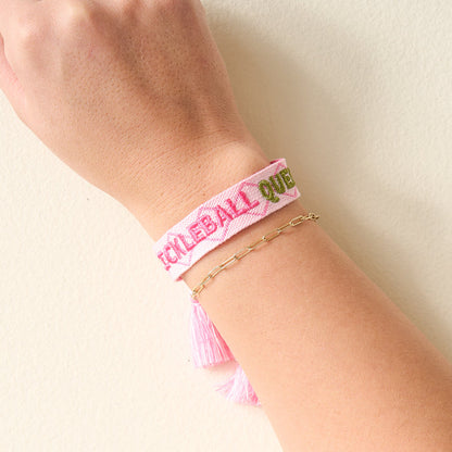 A stylish wrist features The Darling Effect’s Pickleball Queen Woven Bracelet, embroidered with “PICKLEBALL QUEEN” in pink and green, paired with a gold chain and set against a light background.