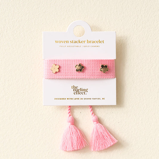 The Darling Effect Pink Woven Bracelet features gold flower charms and two pink tassels, displayed on a white card. Perfect for stacking with your favorite adjustable bracelets.