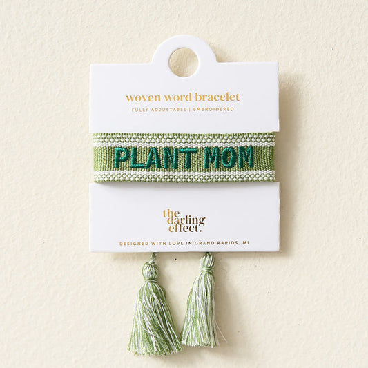The Darling Effect’s Plant Mom Woven Bracelet is a green bracelet with “PLANT MOM” embroidered in teal, tassel ends, and comes on a white card. It’s part of their Adjustable Woven Word Bracelets collection.