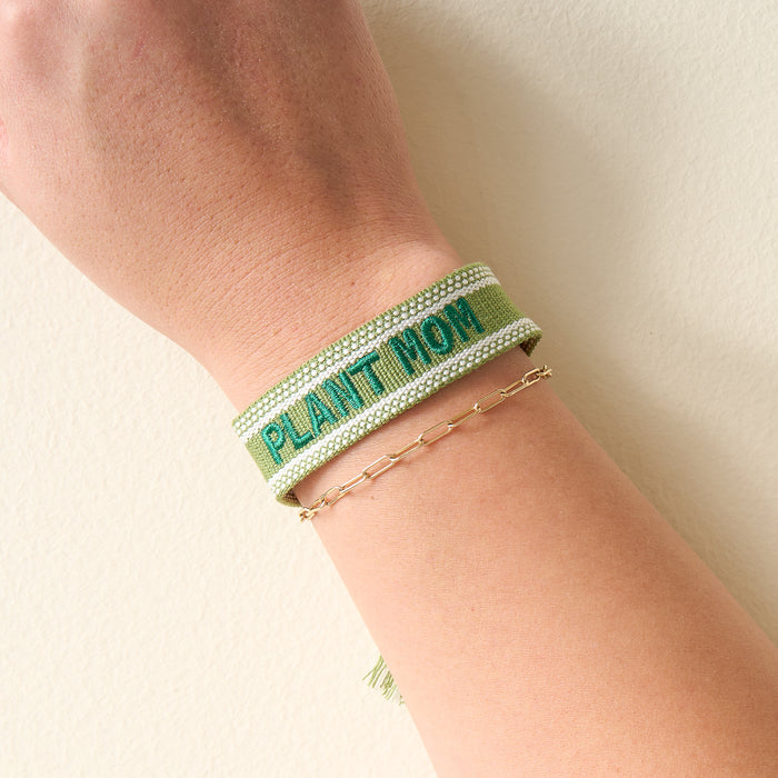 A person’s wrist adorned with The Darling Effect’s Plant Mom Woven Bracelet in green and a delicate gold beaded chain—stylish accessories highlighted on a pale, neutral background.