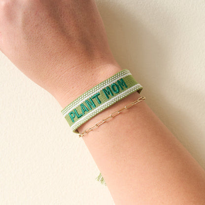 A person’s wrist adorned with The Darling Effect’s Plant Mom Woven Bracelet in green and a delicate gold beaded chain—stylish accessories highlighted on a pale, neutral background.