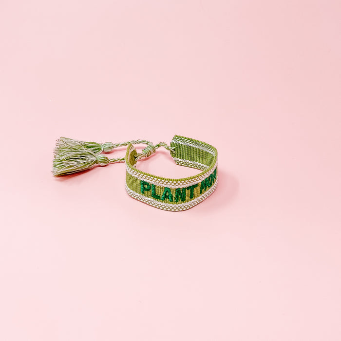 The Darling Effect's Plant Mom Woven Bracelet features "PLANT" in green on a green and white adjustable band with tassels, shown on a pastel pink background. Ideal for trendy accessory lovers and fans of unique embroidered bracelets.