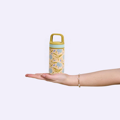 A hand with a delicate bracelet holds The Darling Effect's Poppin' Petals Aqua 6 oz Lil' Bitty Bottle in pastel yellow, featuring floral and bird designs—an ideal travel companion for hydration on the go against a plain light background.