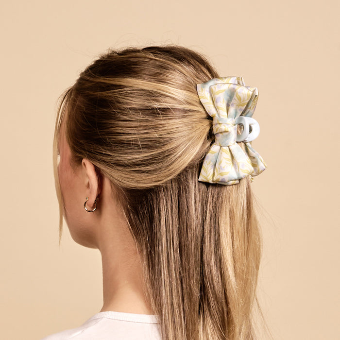 A woman with long, straight blonde hair is shown from the back, wearing The Darling Effect’s Poppin' Petals Aqua Mini Satin Bow Claw Clip and small hoop earrings against a plain beige background.