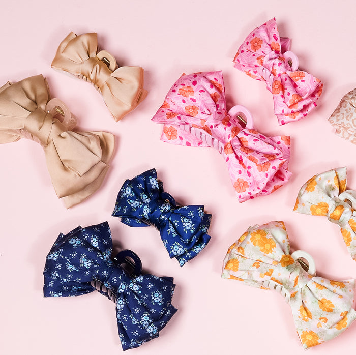The Darling Effect’s Poppin' Petals Aqua Mini Satin Bow Claw Clip is featured among charming assorted hair bows in beige, pink, navy blue, and white with floral patterns on a light pink background.