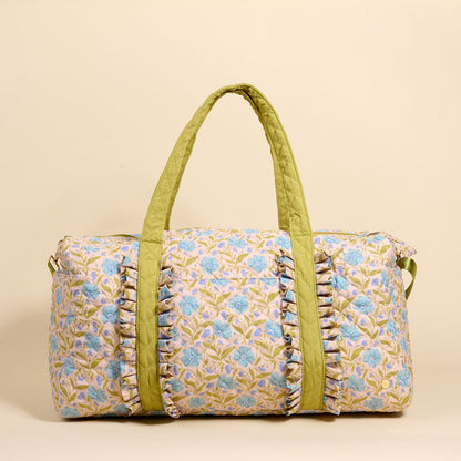 The Darling Effect’s Poppin' Petals Aqua On-the-Go Quilted Weekender Bag features a blue and green floral pattern, light olive green handles, and front ruffle details—ideal for travel. Shown against a beige background.