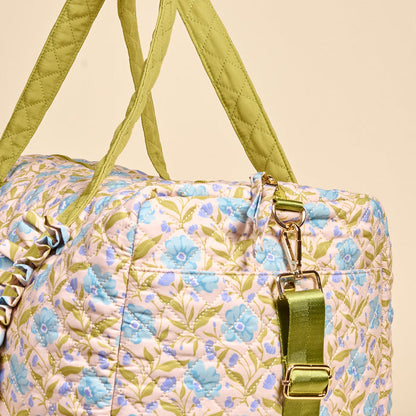 The Darling Effect's Poppin' Petals Aqua On-the-Go Quilted Weekender Bag, featuring a blue floral pattern, green straps, gold hardware, and an adjustable shoulder strap, is shown against a beige background.