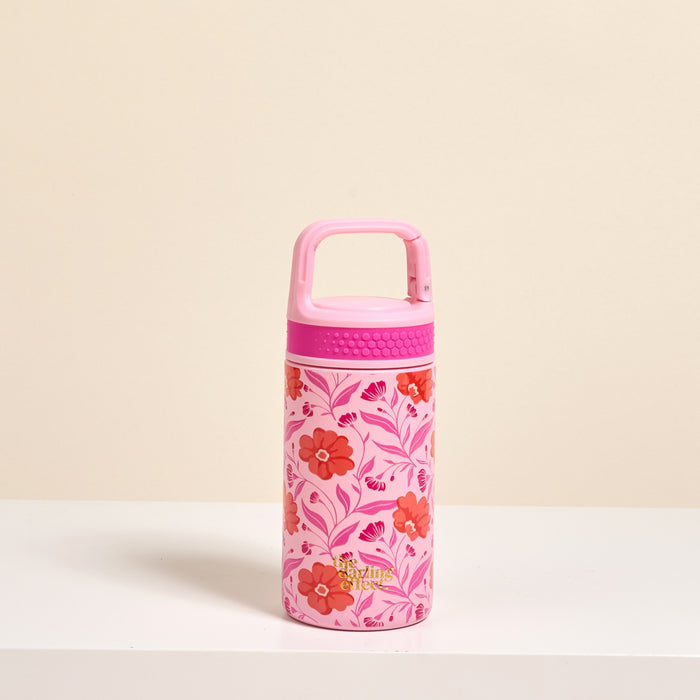 The Darling Effect’s Poppin’ Petals Hot Pink 6 oz Lil’ Bitty Bottle, featuring a handle lid and red-pink floral design, sits on a white surface against a beige background—your ideal stylish travel companion.