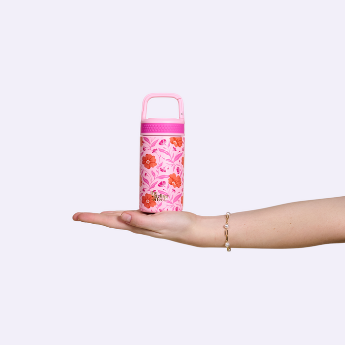 A hand adorned with a delicate bracelet holds the Poppin' Petals Hot Pink 6 oz Lil' Bitty Bottle by The Darling Effect, its floral design making it a charming travel companion against a light background.