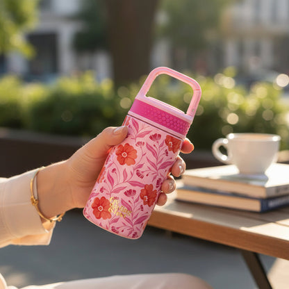 A person holds The Darling Effect’s Poppin' Petals Hot Pink 6 oz Lil' Bitty Bottle, featuring a pink handle and red floral pattern, at an outdoor table with books and a coffee cup—the perfect companion for any adventure.